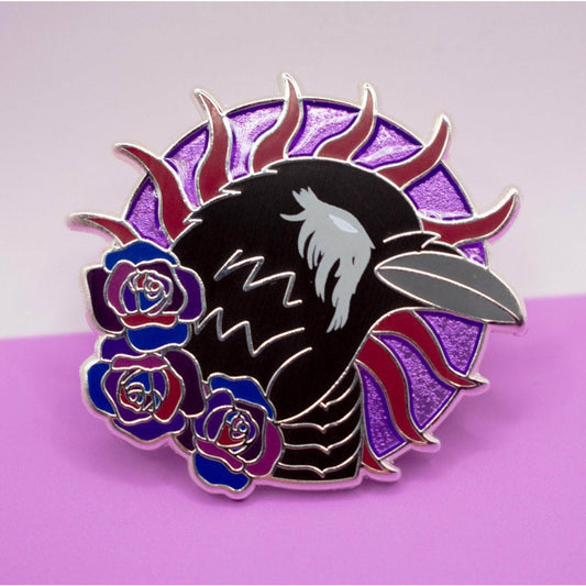 In the photo is a silver metal pin featuring a black crow looking right. The crow has a grey beak and a white eye with grey feathers around it. The crow is on a round purple background with violet spokes going around it. The round purple background is transluscent enamel to look 3D. Beneath the crow at its neck are three roses in various shades of purple.