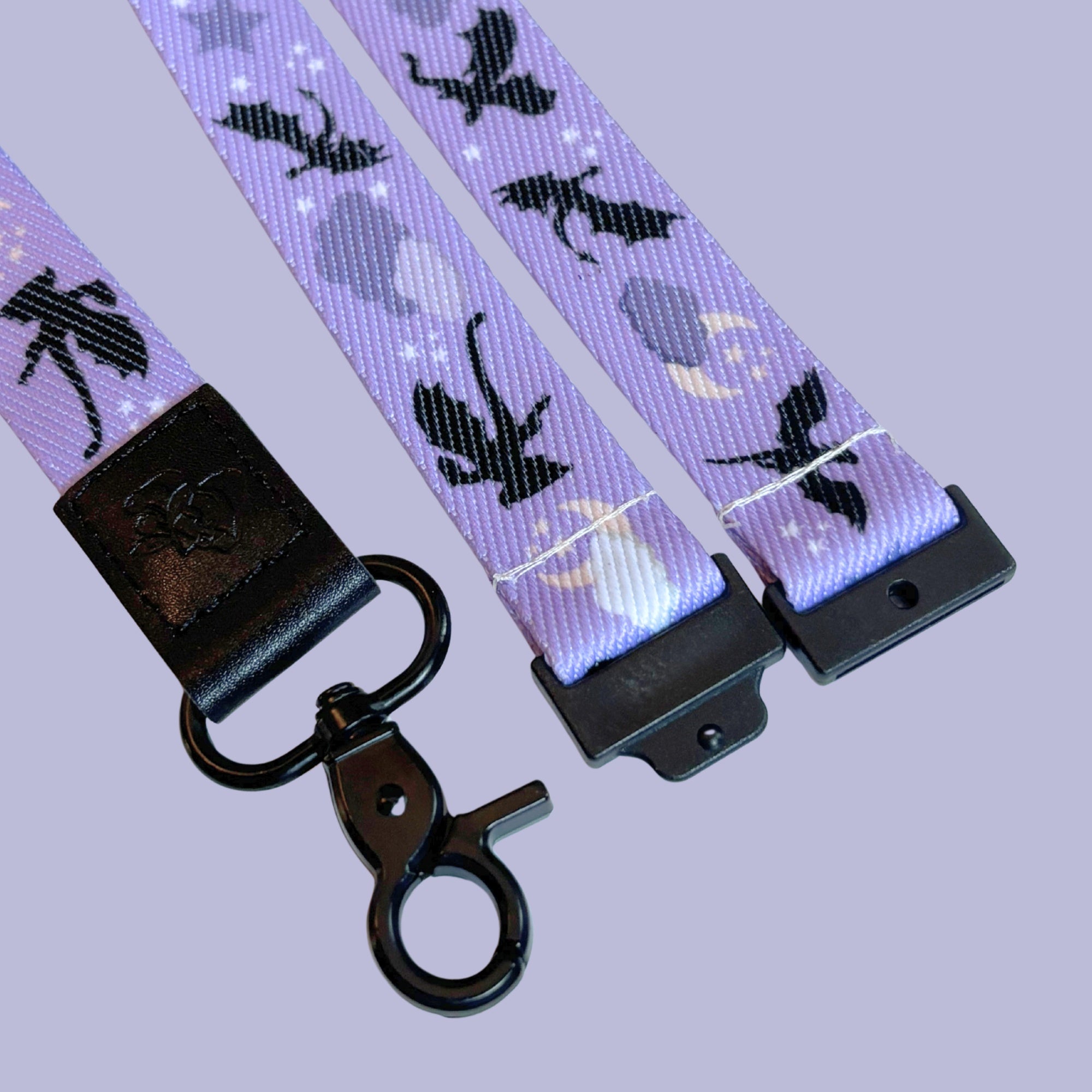 A close-up of a purple lanyard featuring a repeating pattern of black silhouettes of dragons, together with moons, stars, and clouds, finished with a black breakaway accessory and a black metal clasp.