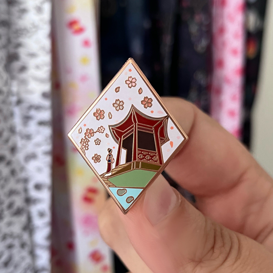Enamel pin with a design of a traditional building and cherry blossoms held by a hand against a colorful background.