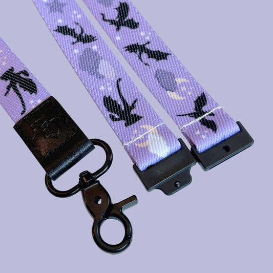A close-up of a purple lanyard featuring a repeating pattern of black silhouettes of dragons, together with moons, stars, and clouds, finished with a black breakaway accessory and a black metal clasp.