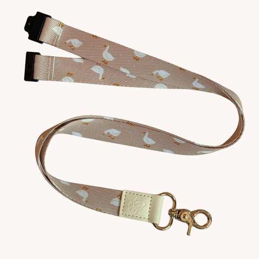 A fabric lanyard strap with a repeating white goose pattern on a light beige background, featuring black plastic quick-release safety buckle on one end and a gold-tone metal swivel clasp with a cream-colored leather patch on the other.